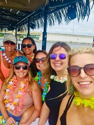 Six smiling women wearing sunglasses and colorful leis on a tiki-style boat near a waterfront bridge, lively daytime coastal boat party.
