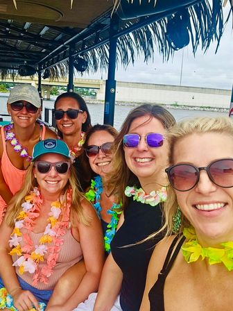Six smiling women wearing sunglasses and colorful leis on a tiki-style boat near a waterfront bridge, lively daytime coastal boat party.