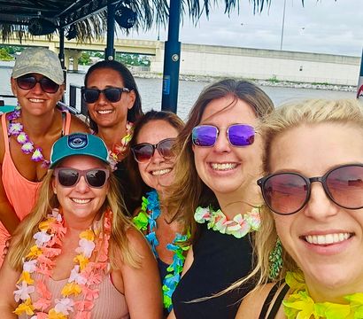 Six smiling women wearing sunglasses and colorful leis on a tiki-style boat near a waterfront bridge, lively daytime coastal boat party.