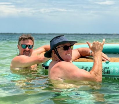 Two men in sunglasses and a sun hat smiling and relaxing in clear shallow coastal water beside a turquoise inflatable paddleboard, one giving a friendly shaka gesture under a blue sky — sunny beach day.
