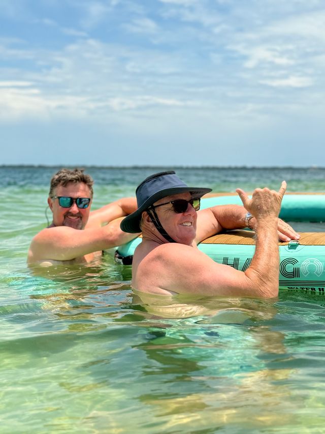 Two men in sunglasses and a sun hat smiling and relaxing in clear shallow coastal water beside a turquoise inflatable paddleboard, one giving a friendly shaka gesture under a blue sky — sunny beach day.