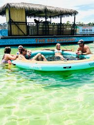 Group of friends lounging on a round inflatable floating lounge in clear turquoise shallow water next to a tiki-style party boat with a thatched roof — sunny beach vibes.