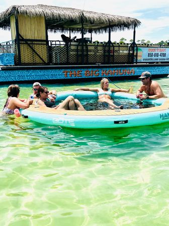 Group of friends lounging on a round inflatable floating lounge in clear turquoise shallow water next to a tiki-style party boat with a thatched roof — sunny beach vibes.