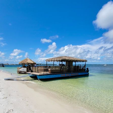 Tiki-style pontoon boat with thatched roofs moored on a white sandy beach in clear turquoise shallow water beneath a bright blue sky with fluffy clouds.