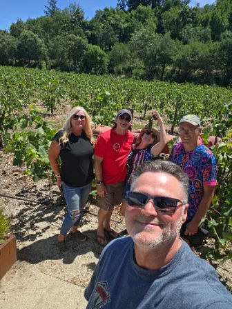 Group selfie of five adults smiling in a sunny wine-country vineyard, standing among rows of green grapevines with oak-covered hills and blue sky in the background.