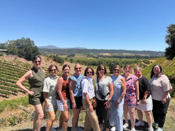 Eleven women smiling and posing at a sunny vineyard overlook with rows of grapevines, rolling wine-country hills and a clear blue sky.