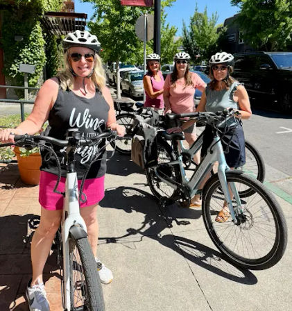 Four women wearing helmets with e-bikes on a sunny tree-lined downtown sidewalk, smiling and ready for a summer bike ride.