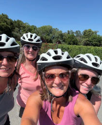 Four women wearing white bike helmets and sunglasses smiling for a selfie on a sunny bike path with green trees and a clear blue sky