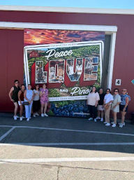 Group of visitors posing in front of a large colorful postcard-style mural reading “Peace Love and Pies” with vineyard and blueberry imagery on a sunny day in an outdoor parking area