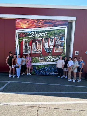 Group of visitors posing in front of a large colorful postcard-style mural reading “Peace Love and Pies” with vineyard and blueberry imagery on a sunny day in an outdoor parking area