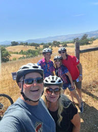 Group selfie of five helmeted cyclists smiling on a sunny rural bike ride, posed by a wooden fence with golden rolling hills and a clear blue sky in the background.