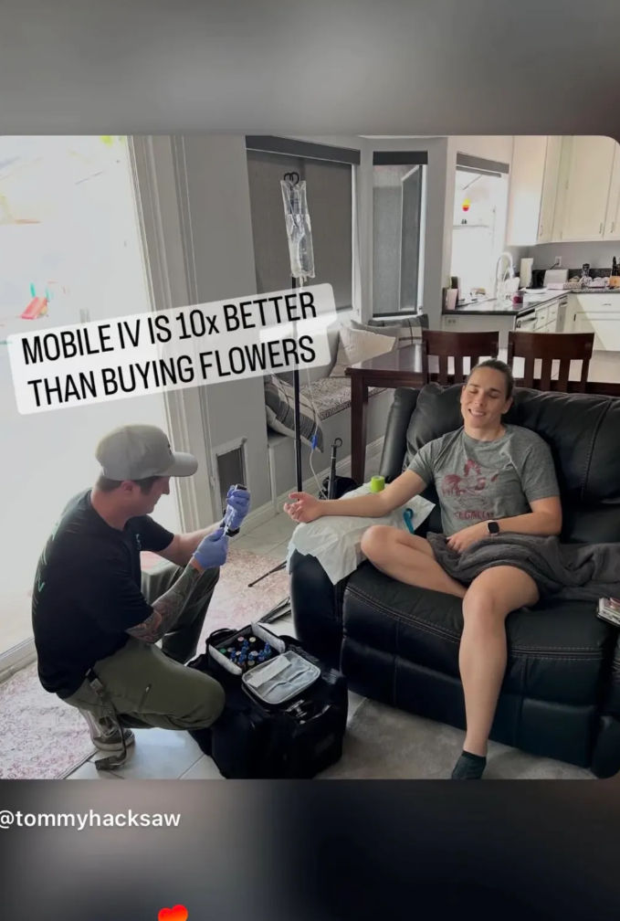 Person relaxing on a living room recliner receiving mobile IV drip from a technician kneeling with a medical kit — in-home IV therapy and wellness service near the kitchen.
