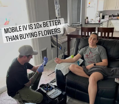 Person relaxing on a living room recliner receiving mobile IV drip from a technician kneeling with a medical kit — in-home IV therapy and wellness service near the kitchen.