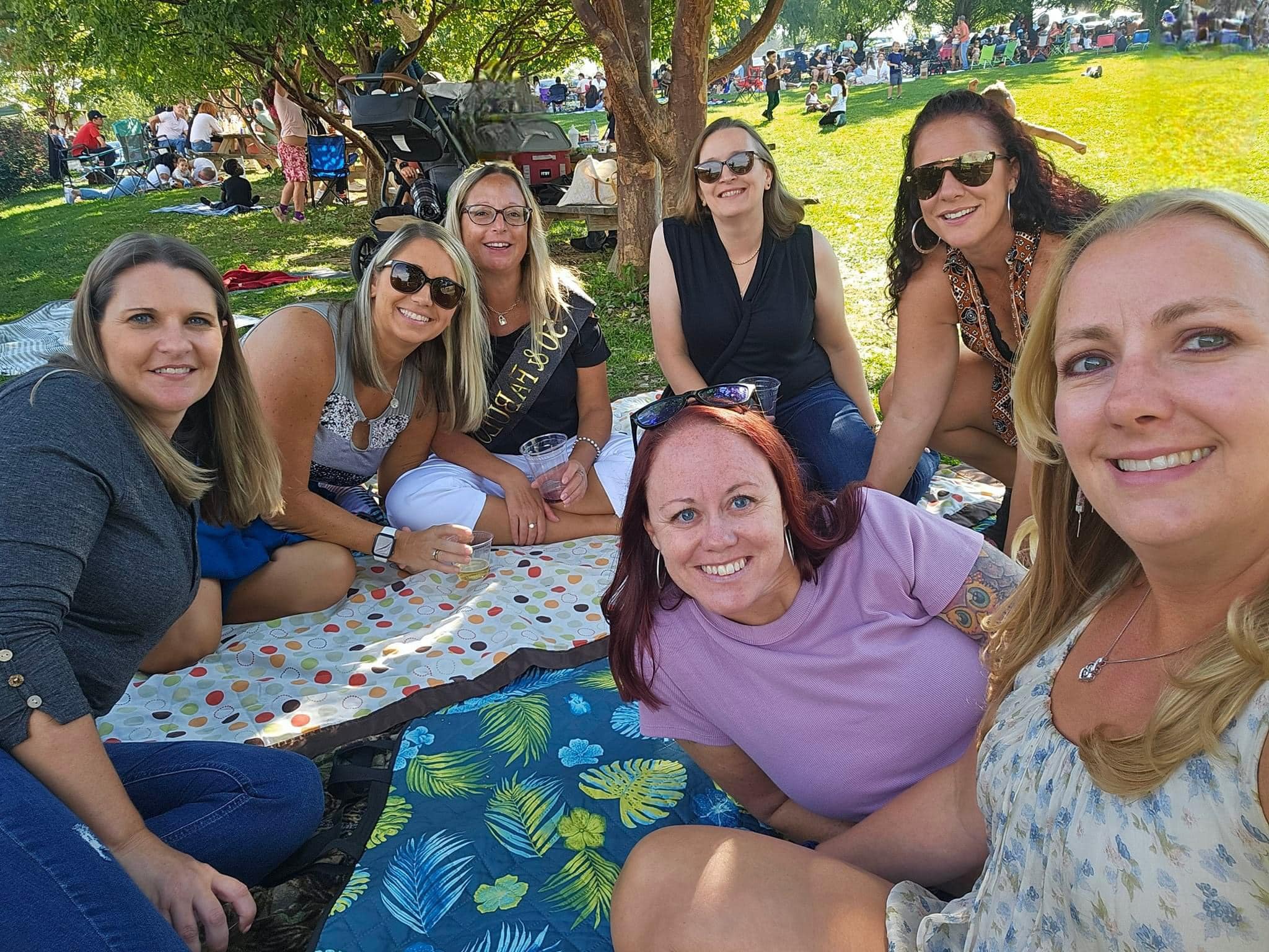 Seven women smiling on picnic blankets under trees at a sunny park, enjoying drinks at an outdoor festival