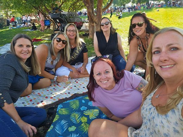 Seven women smiling on picnic blankets under trees at a sunny park, enjoying drinks at an outdoor festival