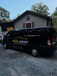 Sleek black passenger shuttle van with yellow transport logo parked on a gravel driveway in front of a single-story suburban house at dusk, framed by trees.