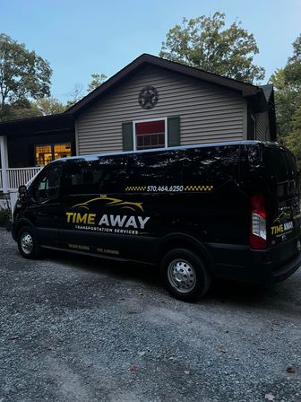 Sleek black passenger shuttle van with yellow transport logo parked on a gravel driveway in front of a single-story suburban house at dusk, framed by trees.