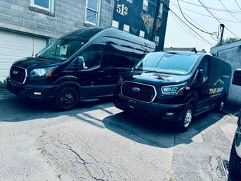 Two sleek black Ford Transit cargo vans parked side-by-side on cracked pavement beside a cinderblock building with a painted mural and overhead utility wires in an urban alley