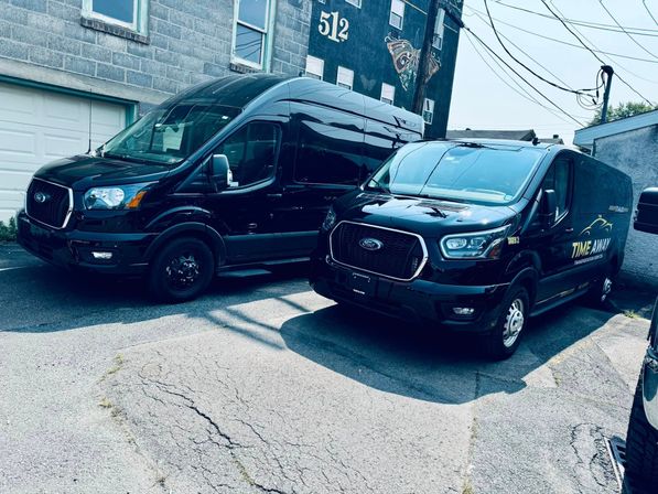 Two sleek black Ford Transit cargo vans parked side-by-side on cracked pavement beside a cinderblock building with a painted mural and overhead utility wires in an urban alley