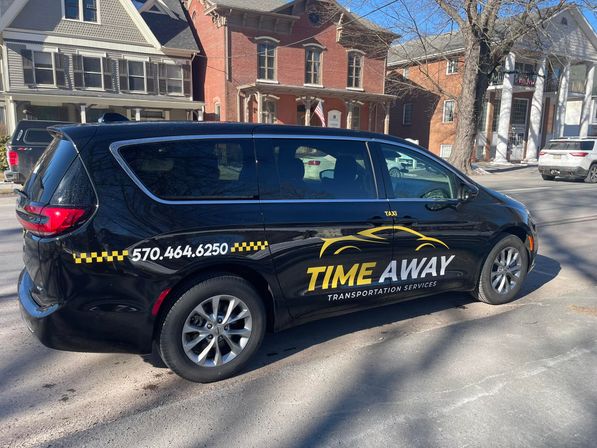 Side view of a black taxi minivan with yellow checkered stripe and stylized car graphic parked on a sunny residential street lined with historic brick and wood-frame houses