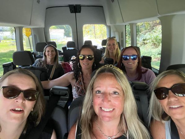 Seven smiling women wearing sunglasses taking a selfie inside a passenger van on a sunny countryside road trip.