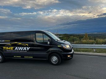 Black transport van with yellow-and-white logo parked by a roadside guardrail at a hilltop scenic overlook, dramatic cloudy sunset sky and rolling green landscape in the background.