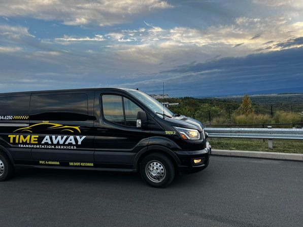 Black transport van with yellow-and-white logo parked by a roadside guardrail at a hilltop scenic overlook, dramatic cloudy sunset sky and rolling green landscape in the background.