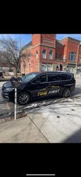 Black passenger van with yellow-and-white transportation logo parked at a downtown curb by parking meters, in front of a red-brick City Hall on a small-town main street with traces of snow on the sidewalk.