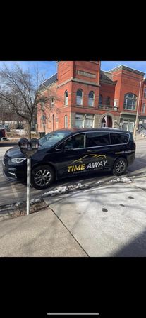 Black passenger van with yellow-and-white transportation logo parked at a downtown curb by parking meters, in front of a red-brick City Hall on a small-town main street with traces of snow on the sidewalk.