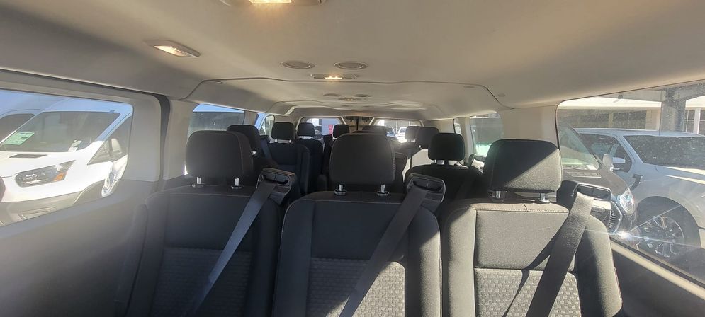 Spacious passenger van interior with multiple rows of black cloth seats and seat belts, overhead lights and daylight streaming through side windows while parked in a vehicle lot.