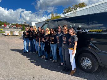 Smiling women in matching black T‑shirts with colorful tree graphic lined up beside a black passenger van in a sunny fall gravel lot with pumpkins, blue sky, and fluffy clouds.