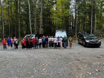Group of young people lined up in a wooded parking area with two black vans and a black SUV, tall trees and scattered autumn leaves in the background.