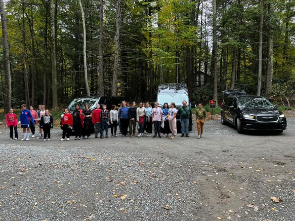 Group of young people lined up in a wooded parking area with two black vans and a black SUV, tall trees and scattered autumn leaves in the background.