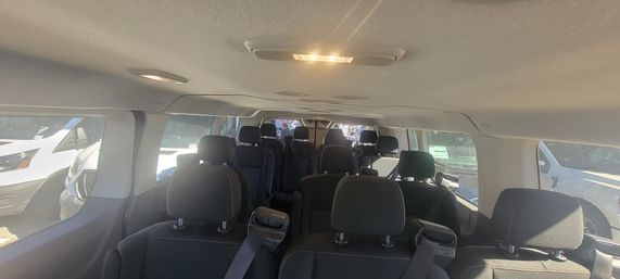 Spacious multi-row passenger van interior with black cloth seats, headrests and seatbelts, overhead dome light on and sunlight streaming through side windows showing other vans in a parking lot.