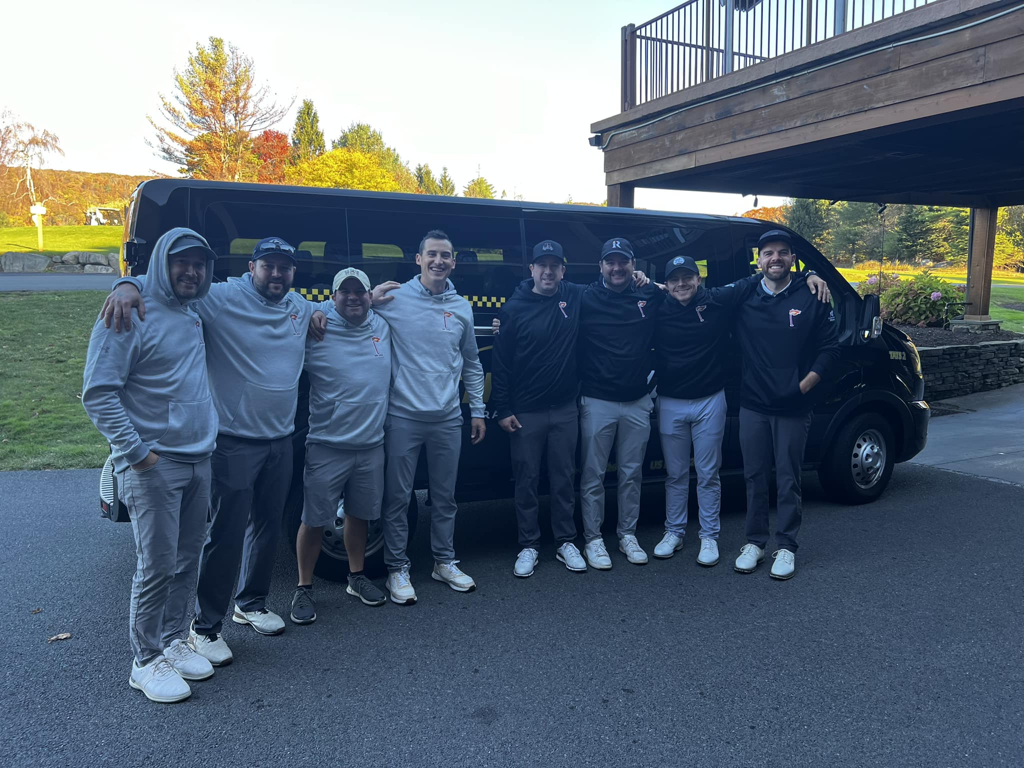 Cheerful group in matching golf apparel posing arm-in-arm in front of a black shuttle van at a golf course with autumn trees and a clubhouse deck in the background
