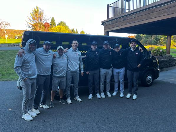 Cheerful group in matching golf apparel posing arm-in-arm in front of a black shuttle van at a golf course with autumn trees and a clubhouse deck in the background