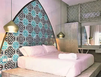 Chic Mediterranean-style boutique bedroom with raised platform king bed in blush linens, turquoise patterned arched tile headboard, brass pendant lights, wooden wardrobe and vanity mirror.