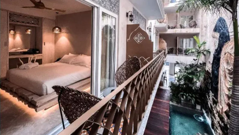 Balcony view of a tropical boutique hotel: sliding-door luxury room with low platform bed, hanging wicker chairs and decorative railing overlooking a lush indoor courtyard with palms and a narrow pool with waterfall.