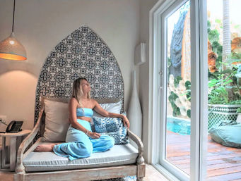 Person in a turquoise lounge set relaxing on a carved wooden daybed under a patterned tile arch, gazing through sliding glass doors at a lush poolside patio with tropical plants.