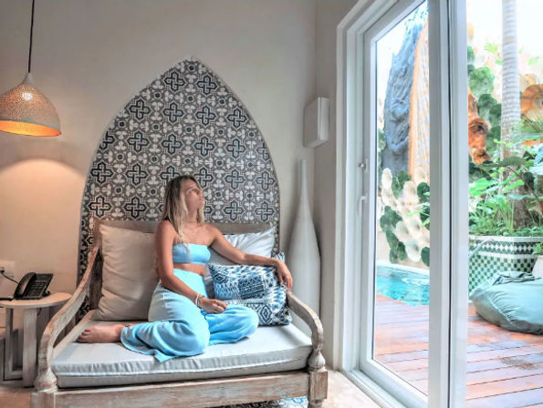 Person in a turquoise lounge set relaxing on a carved wooden daybed under a patterned tile arch, gazing through sliding glass doors at a lush poolside patio with tropical plants.