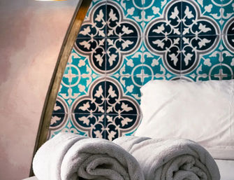 Two rolled white towels on a bed with a pillow, set against an ornate teal-and-navy patterned tile headboard in a cozy bedroom.