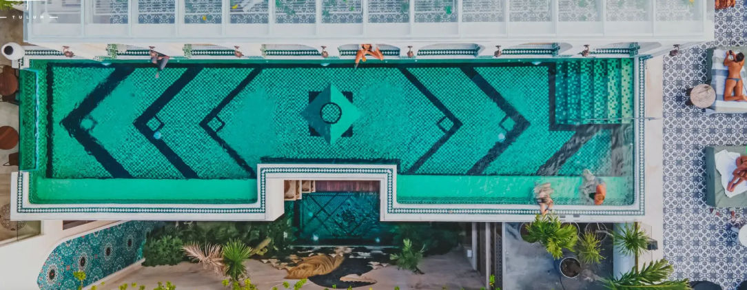 Aerial view of a turquoise mosaic rooftop pool with bold geometric patterns, sunbathers on lounge chairs, and patterned tile deck at a tropical Tulum resort.