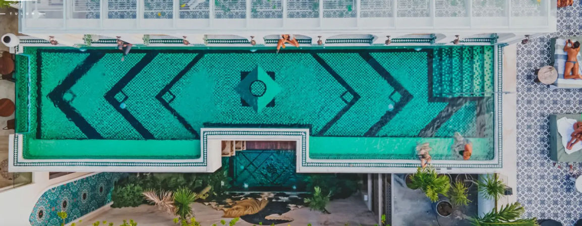 Aerial view of a turquoise mosaic rooftop pool with bold geometric patterns, sunbathers on lounge chairs, and patterned tile deck at a tropical Tulum resort.