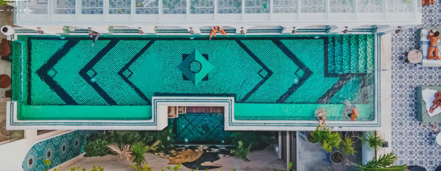Aerial view of a turquoise mosaic rooftop pool with bold geometric patterns, sunbathers on lounge chairs, and patterned tile deck at a tropical Tulum resort.