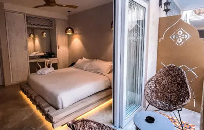 Cozy boutique-style bedroom with illuminated low platform bed, warm pendant lamps and ceiling fan, sliding glass doors to a small balcony featuring a woven rattan nest chair, round table and decorative screen — neutral, ambient-lit retreat.