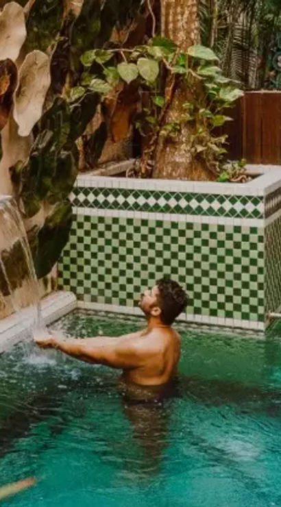 Man enjoying a small outdoor tiled plunge pool with green mosaic tiles and a decorative waterfall wall in a tropical courtyard