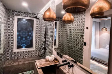 Moroccan-inspired bathroom with black-and-white geometric mosaic tiles, large mirror, warm copper pendant lights, wall-mounted black shower fixtures, concrete sink with wooden slatted bench and rolled towels, and a bedroom visible through the doorway.