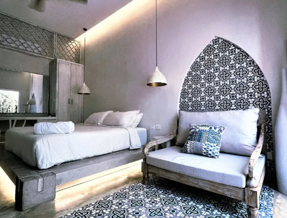 Minimalist bedroom with a raised concrete platform bed in white linens, warm pendant lights, a rustic wooden loveseat with a blue patterned pillow, and a decorative arched black-and-white tile accent that matches the patterned floor.