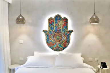 Bright boho bedroom with crisp white bed, glowing colorful Hamsa hand wall art above the headboard, woven pendant lights and minimalist side tables.