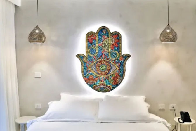 Bright boho bedroom with crisp white bed, glowing colorful Hamsa hand wall art above the headboard, woven pendant lights and minimalist side tables.